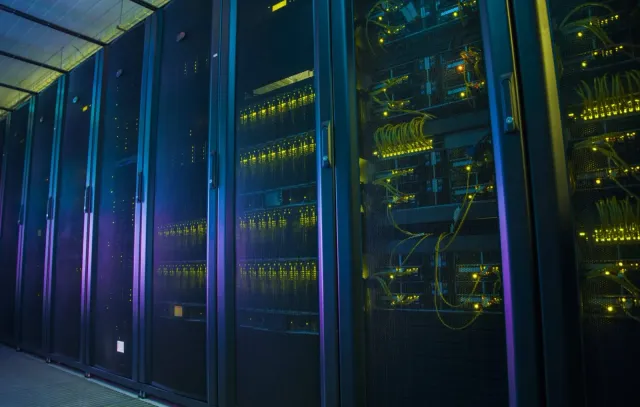 A dimly lit data centre aisle of network racks behind mesh doors, glowing with yellow status LEDs and neat runs of yellow patch cabling.