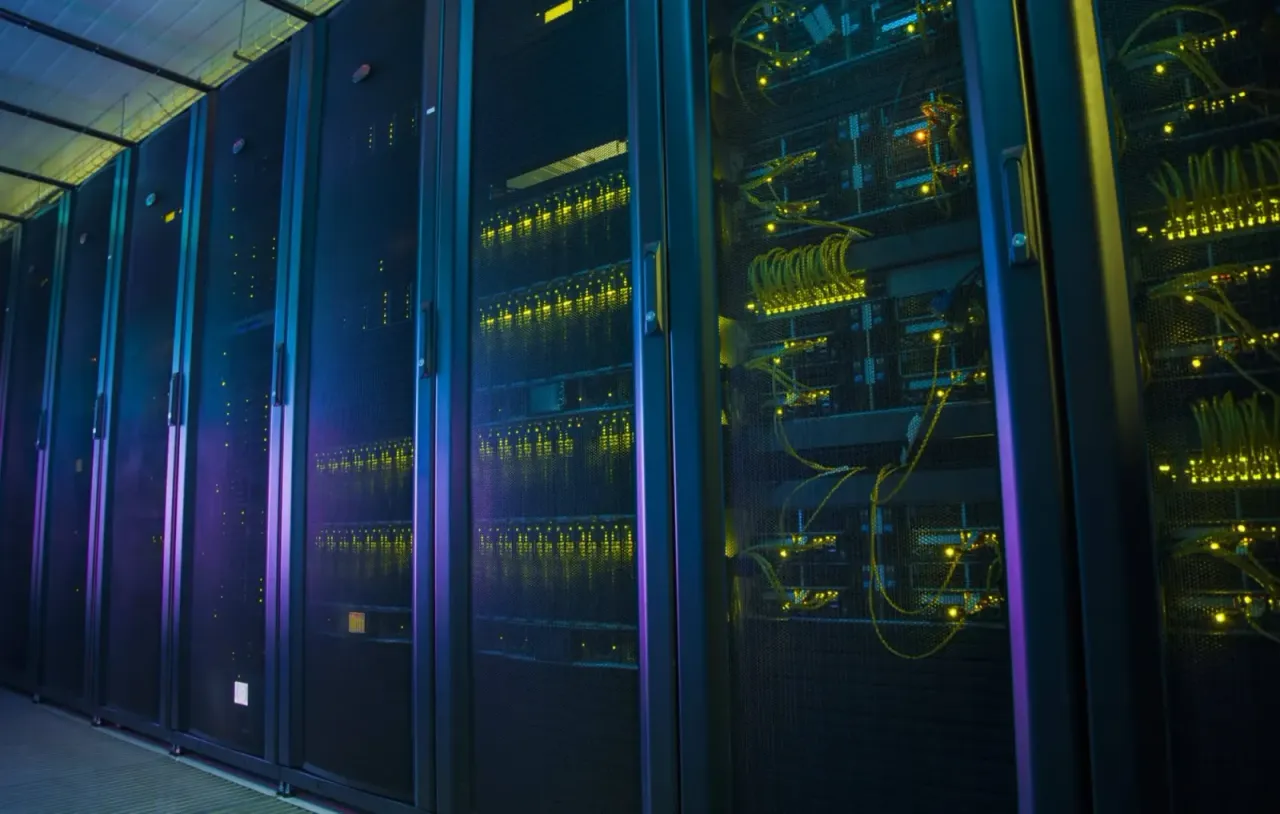 A dimly lit data centre aisle of network racks behind mesh doors, glowing with yellow status LEDs and neat runs of yellow patch cabling.