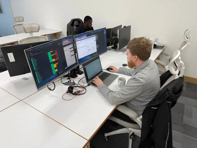 Next2IT engineers at multi-screen workstations, one monitoring a live service dashboard while working on a laptop.